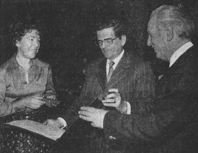 Hutzler Medal of Merit A black-and-white photograph of three people in a dark room. The man in the middle is holding a certificate.