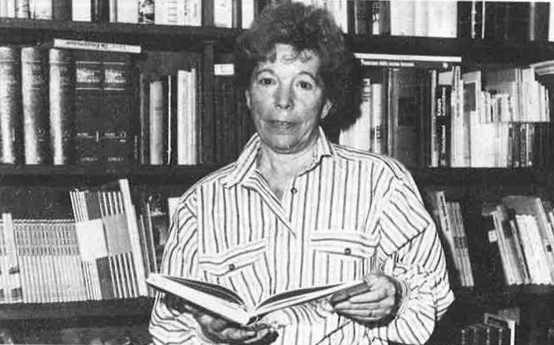 Hutzler Portrait 4 A black-and-white photograph of a woman standing in front of a full bookshelf, holding a book.