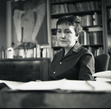 Christa Holscher 1967 Black and white photo of a woman with short hair at a desk in front of a bookshelf.