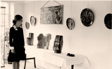 Madeleine Mangold avant une présentation de ses œuvres. Photo en noir et blanc d'une jeune femme debout à côté d'un mur sur lequel a été accroché de l'art céramique.