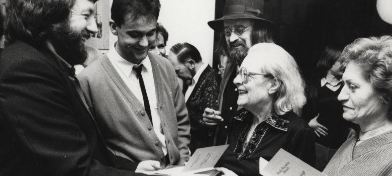 Ruth Ricarda Bruch per la pubblicazione del suo primo volume di poesie. Foto in bianco e nero di Ruth Ricarda Bruch che ride e sta di fronte a due uomini. Uno di loro tiene in mano il suo libro di poesie.