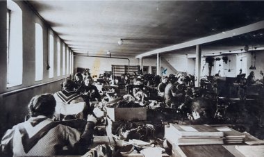 Foto in bianco e nero dell'interno di una fabbrica. Da dietro si vede una fila di operai alle macchine da cucire.