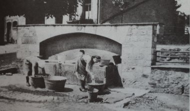 Fotografia in bianco e nero che ritrae due donne in piedi davanti a una fontana con grandi pentole, intente a lavare il bucato.
