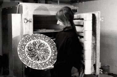 Historyczne zdjęcie Madeleine Mangold i jej pracy. Czarno-białe zdjęcie kobiety przed piecem. Trzyma ozdobny duży talerz skierowany w stronę aparatu.