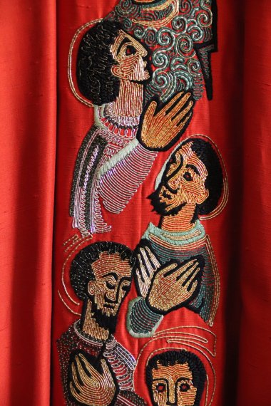 Apostelgewand der Pfarrei Maria vom Frieden Rotes Gewand mit bunten Stickereien, die die Köpfe und Oberkörper von Männern zeigen.