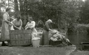 Waschplatz Herbitzheim Schwarz-Weiß-Foto von vier Frauen, die neben einem Fluss stehen und ihre Kleidung in einem Großen Fass waschen.