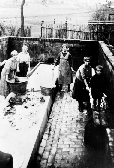 Schwarz-Weiß-Foto von vier Frauen und einem Kind innerhalb eines eingemauerten Bereichs, in dessen Mitte ein flacher Brunnen steht.