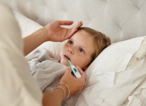 Ein Kind liegt im Bett und die Mutter misst die Temperatur mit Hand und Thermometer