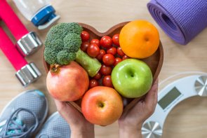 Prävention und Gesundheitsförderung Eine Schüssel in Herzform mit Obst und Gemüse, im Hintergrund sind Fitnessgeräte