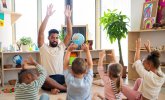 Ein Mann macht sitzend Bewegungsspiele mit Kindern, dabei heben sie die Arme in die Luft