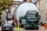 Ein Schwertransport fährt durch eine enge Straße in einem Ort, unterstützt durch einen Lotsen.